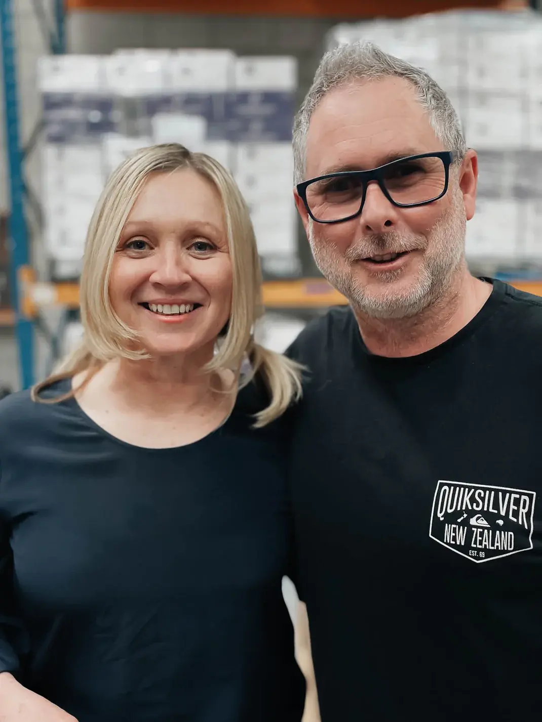 Smiling couple in Molly Woppy warehouse, with stacked boxes in background. Hayley Molloy with blonde hair in pigtails, Alistair Parker with gray hair, beard, and black glasses. Casual clothing, industrial storage environment.
